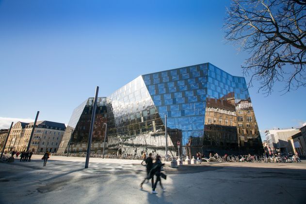 Das Bild zeigt das neue Gebäude vom Platz der Alten Synagoge fotografiert und mit einer gläsernen, verschieden geneigten Fassade. Der Himmel ist blau. Auf der Nordseite spiegelt sich das Stadttheater in der Fassade. Im Vordergrund laufen Menschen. 