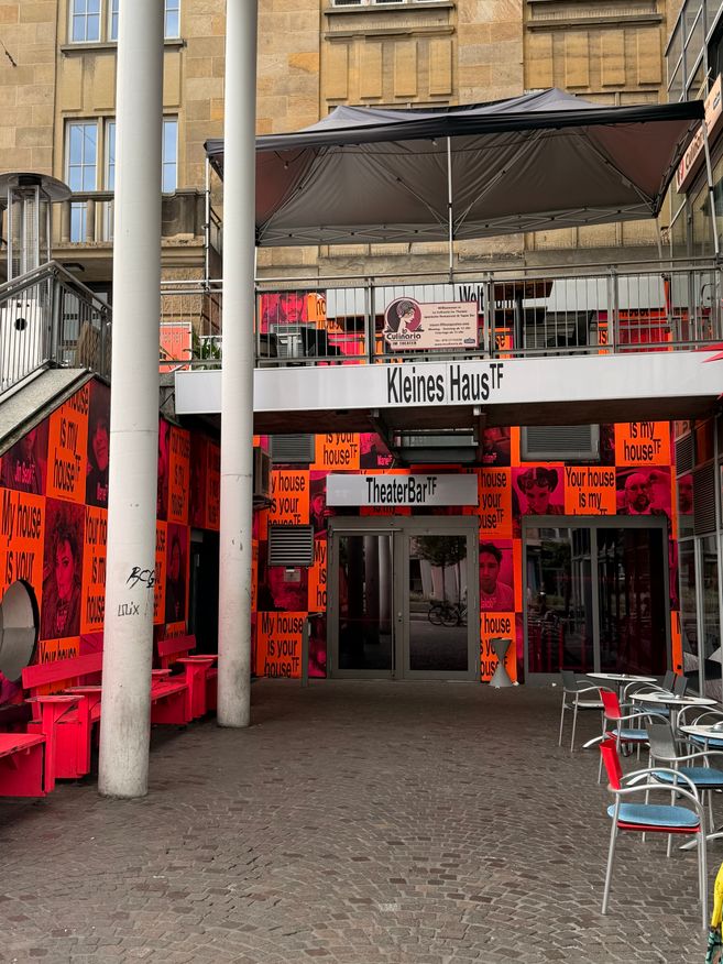 Eingang des TheaterBar im Kleinen Haus des Theaters Freiburg mit bunten Plakaten, Sitzbereich und weißem Sonnendach.