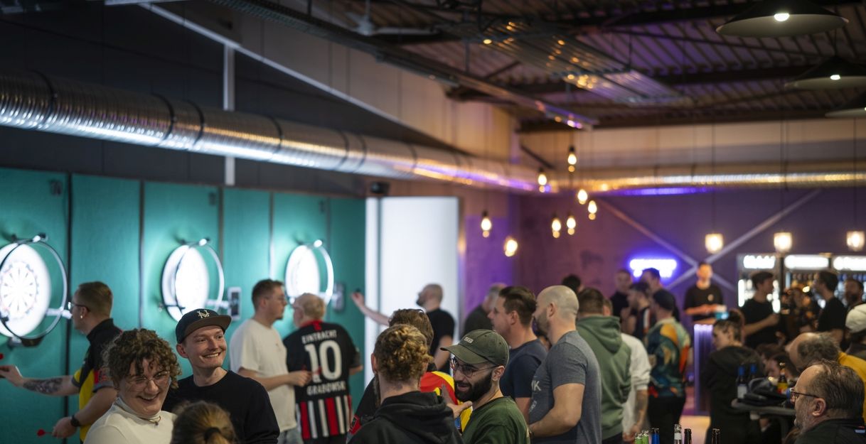 Dartzentrum Freiburg, Dartboards mit Beleuchtung an den Wänden, Bar und Lounge mit warmer Beleuchtung, Menschen, die Darts werfen, zuschauen oder miteinander sprechen. 