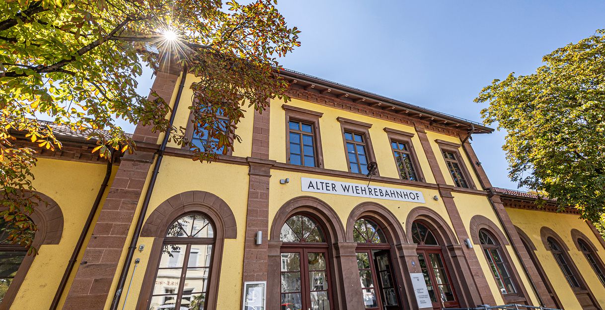 Die Frontansicht des Alten Wiehrebahnhofs mit Bäumen und blauem Himmel