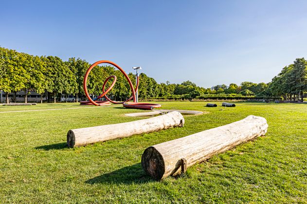 Park mit einem Kunstobjekt in Form eines Gartenschlauches in der Mitte des Parks. Rund um den Schlauch befinden Sie Baumstämme, die Sitzgelegenheiten bieten