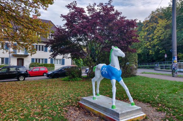 Bunte Pferdeskulptur in einem Park in Freiburg, umgeben von Bäumen und einem Wohnhaus im Hintergrund.