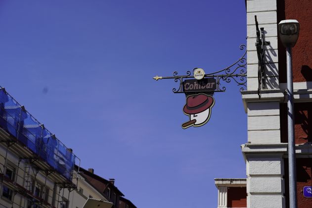 Ein Schild mit der Aufschrift 'Cohibar' und einem stilisierten Gesicht mit Hut und Zigarre hängt an einer Hauswand.