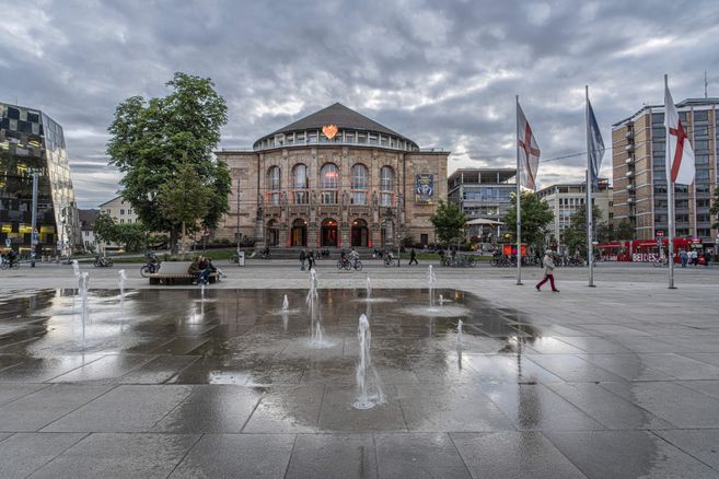 Das Freiburger Stadttheater mit Springbrunnen im Vordergrund und bewölktem Himmel.