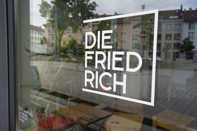 Fensterscheibe mit dem Schriftzug 'DIE FRIEDRICH' in einem städtischen Umfeld.