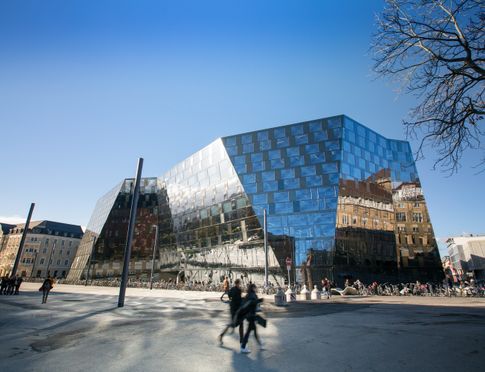 Neues Gebäude der Universitätsbibliothek Freiburg fotografiert vom Platz der Alten Synagoge. Das Gebäude hat eine gläserne, verschieden geneigte Fassade. An der Nordseite spiegelt sich das Stadttheater in der Fassade.