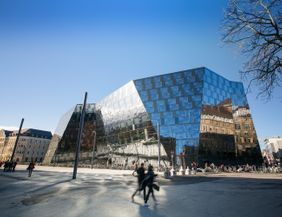 Das Bild zeigt das neue Gebäude vom Platz der Alten Synagoge fotografiert und mit einer gläsernen, verschieden geneigten Fassade. Der Himmel ist blau. Auf der Nordseite spiegelt sich das Stadttheater in der Fassade. Im Vordergrund laufen Menschen. 