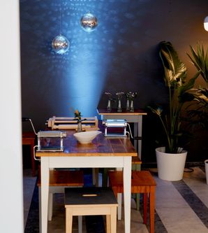 Innenansicht einer stilvollen Food-Boutique mit Holztischen, Pflanzen und hängenden Discokugeln an einer blau beleuchteten Wand.