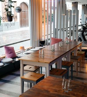 Innenansicht eines modernen Cafés mit Holztischen und Hockern, dekoriert mit Kissen und Pflanzen. Große Fenster bieten Blick auf die Straße.