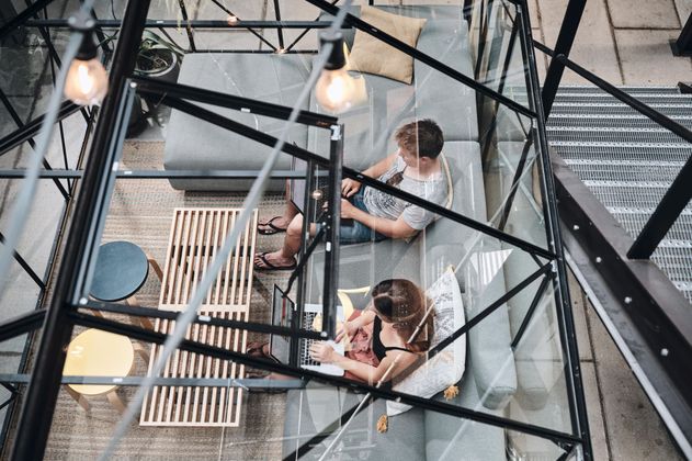 Zwei Personen arbeiten an Laptops in einem modernen, verglasten Raum mit Sofas und Tischen.